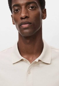 Camisa polo beige con cuello abotonado, de textura suave y sutil detalle de costura, llevada por una persona con un fondo neutro.