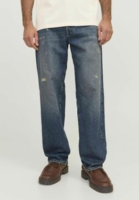 Denim jeans in dark blue with a relaxed fit, featuring a distressed detail on the knee and worn effects, paired with brown leather shoes.