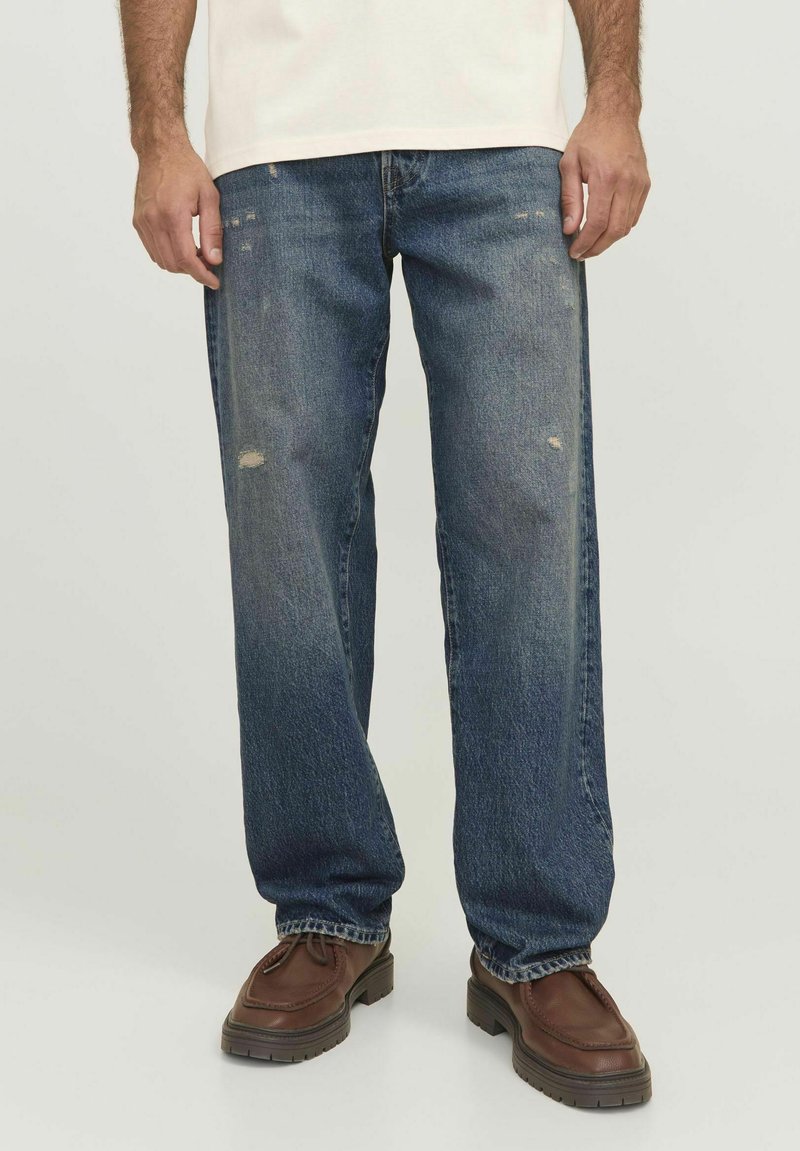 Denim jeans in dark blue with a relaxed fit, featuring a distressed detail on the knee and worn effects, paired with brown leather shoes.