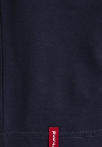 Zbliżenie na granatową tkaninę z małą czerwoną metką z białym napisem „hummel” w pobliżu obszytego brzegu.