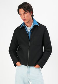 Young man with dark hair wearing a black zip-up jacket over a denim shirt and striped polo, hands in light blue jeans pockets, looking away.