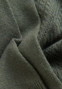 Närbild av mörkgrön stickad tyg med vikta, texturerade och ribbade sektioner som visar stygnsdetaljer.