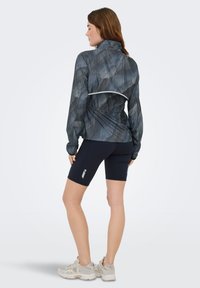 Giacca sportiva leggera in grigio scuro con motivo geometrico, abbinata a pantaloncini aderenti blu navy. Presenta un colletto alto e una vita elastica.