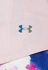 Top deportivo rosa claro hecho de una tela elástica, con un logotipo de Under Armour en degradado azul y verde. Se aprecian detalles de costura.