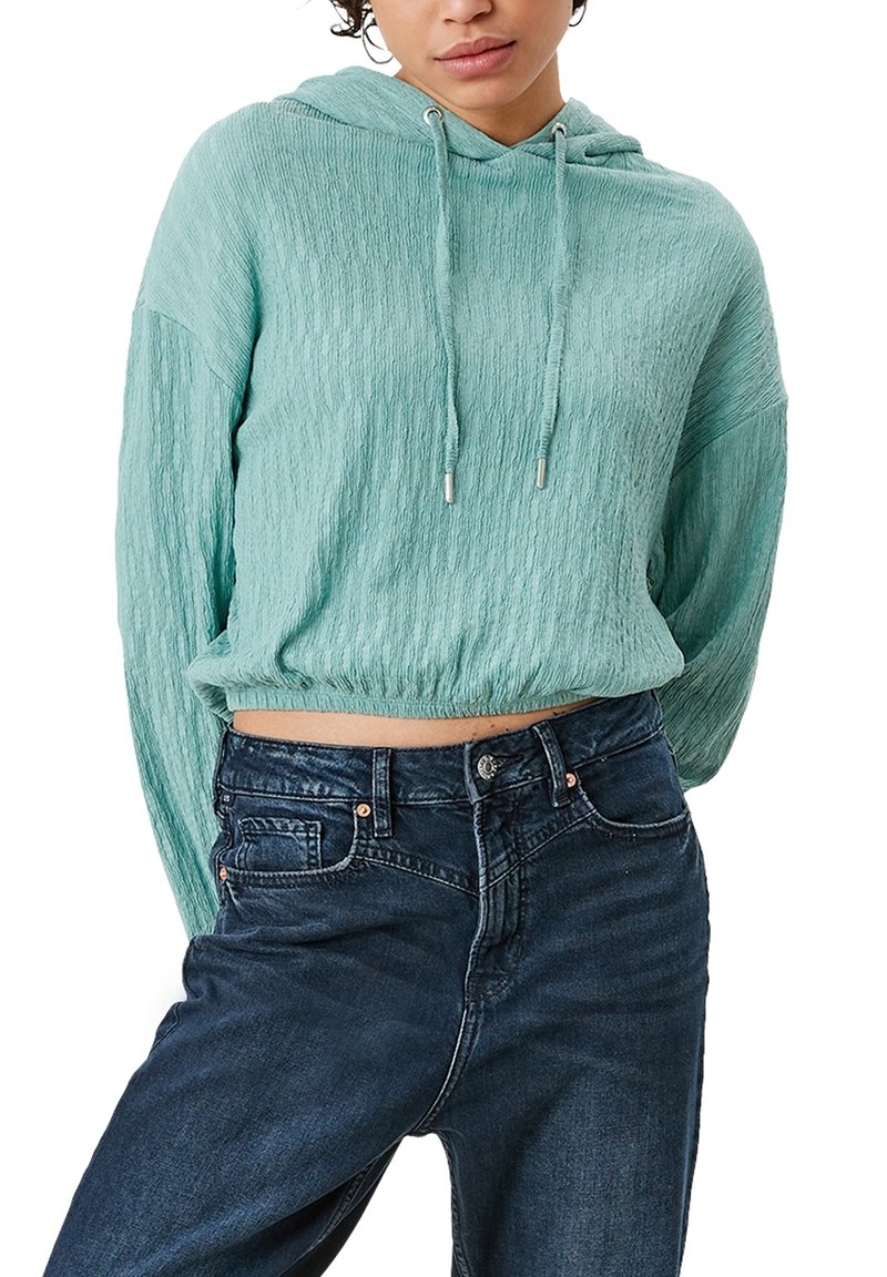 Felpa corta color turchese chiaro con finitura testurizzata, cappuccio con cordoncino e vestibilità casual, abbinata a jeans blu scuro con tasche frontali.