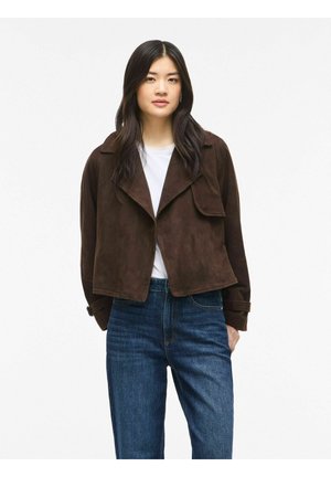 Jeune femme portant une veste courte marron foncé par-dessus une chemise blanche et un jean bleu, debout devant un fond blanc uni.