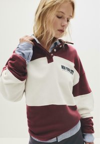 Una donna bionda sistema il colletto di una felpa bordeaux e bianca di New York City sopra una camicia a righe blu, guardando in basso.