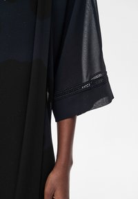 Ciemnoskóra ręka ubrana w czarny strój z szerokim, półprzeźroczystym rękawem ozdobionym delikatną koronką na jednolitym białym tle.