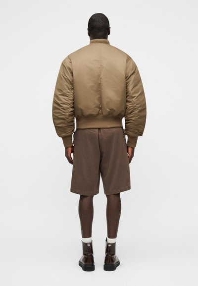 Άνδρας που στέκεται με την πλάτη στραμμένη, φοράει μια μπεζ μπλούζα τύπου μπόμερ, καφέ σορτς μήκους έως το γόνατο, λευκές κάλτσες και σκούρα καφέ μποτάκια σε απλό φόντο.
