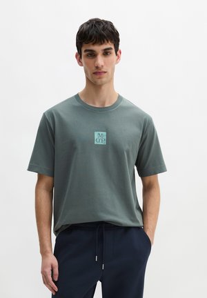 Młody mężczyzna z krótkimi ciemnymi włosami w stonowanym zielonym T-shircie z małym logo „MOP”, stojący na jednolitym jasnym tle.