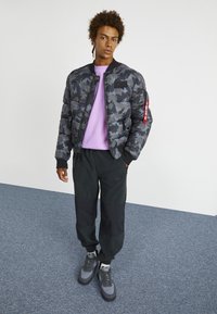 Pletená bomber bunda v černé a šedé kamuflážním vzoru, spárovaná se světle fialovou mikinou, černými kalhotami a šedými teniskami, stojící na šedé podlaze.