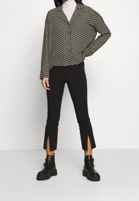 Schwarz-weiß gemusterte Bluse mit Kragen, kombiniert mit schwarzen Hosen mit seitlichen Schlitzen am Saum und klobigen schwarzen Stiefeln.