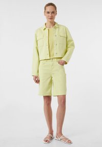 Jasnożółta dżinsowa kurtka z dwiema przednimi kieszeniami, połączona z pasującą żółtą bluzką i szortami. Model nosi białe sandały na ramiączkach.