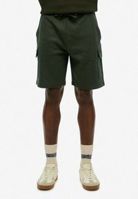 Spodenki cargo w ciemnozielonym materiale z elastycznym pasem, sznurkiem i kieszeniami bocznymi. Noszone z jasnymi sneakersami i skarpetkami.