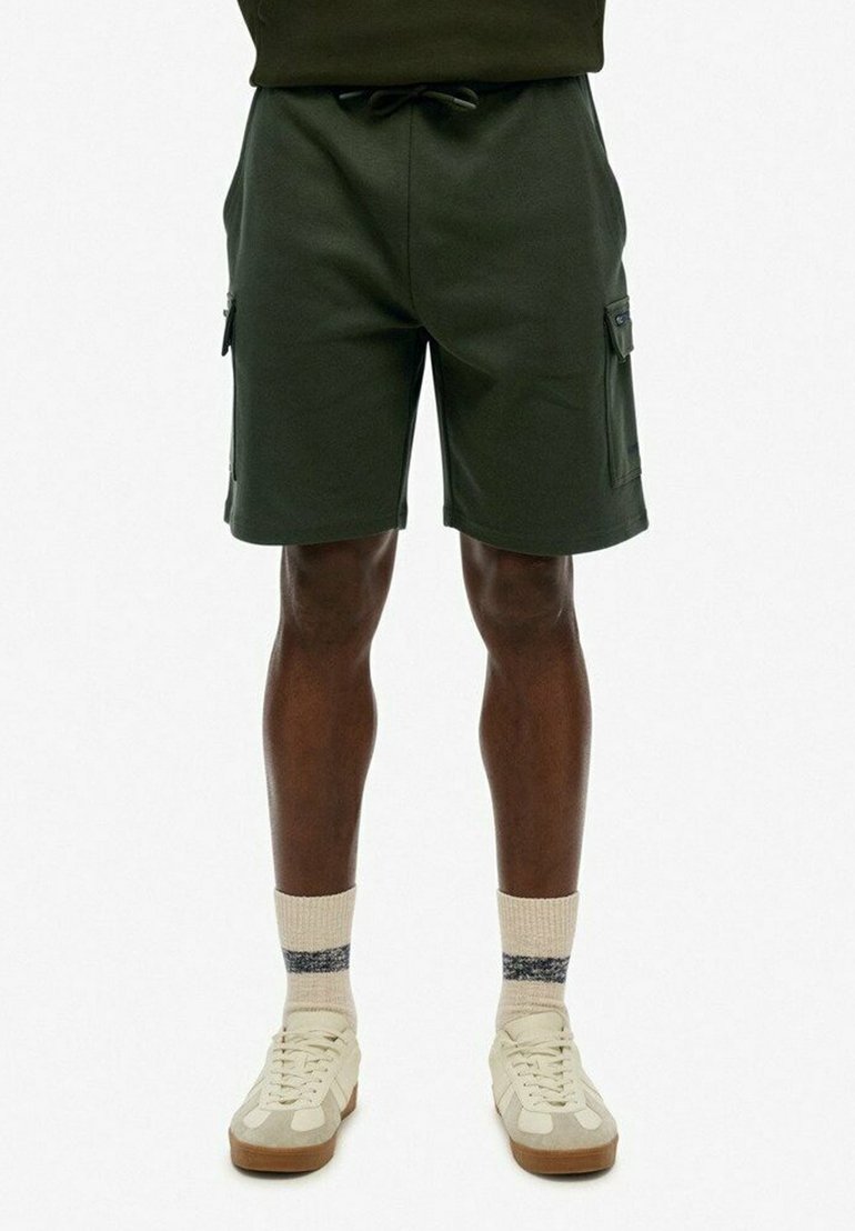 Spodenki cargo w ciemnozielonym materiale z elastycznym pasem, sznurkiem i kieszeniami bocznymi. Noszone z jasnymi sneakersami i skarpetkami.