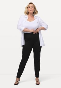 Pantaloni neri a vita alta con una textura liscia, abbinati a una canotta bianca e a una camicia bianca aperta. Tacchi alti neri.
