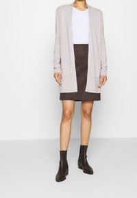 Larga chaqueta de punto color beige claro, con mangas largas, bolsillos frontales, combinada con una falta de ante marrón oscuro y botines negros. Textura suave en toda la prenda.