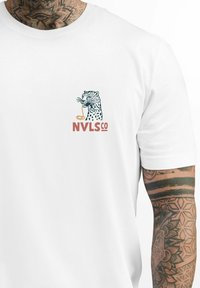Weißes T-Shirt mit kurzen Ärmeln, auf dem ein Leoparden-Graphic mit einem Getränk und dem Text "NVLSCO" in Orange und Blau abgebildet ist. Aus weichem Baumwollstoff.