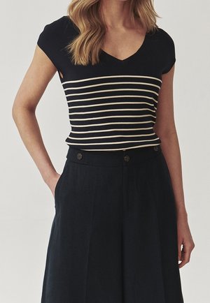 Vrouw draagt een zwart shirt met korte mouwen met witte horizontale strepen en een zwarte hoge taille broek met knoopdetails.