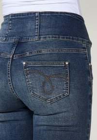 Vaqueros de mezclilla azul de cintura alta con costuras oscuras, que presentan un diseño de bolsillo distintivo con un patrón de lazo decorativo y pequeños acentos de remache.