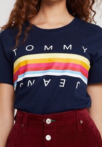 Camiseta de algodón azul marino con gruesas rayas horizontales en amarillo, rosa y azul claro, con "TOMMY JEANS" impreso en blanco en el pecho.