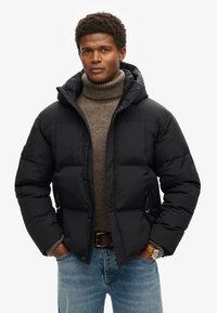 Giacca a vento nera con cappuccio, frontale con zip, tasche laterali e texture trapuntata, indossata sopra un dolcevita marrone e jeans blu.