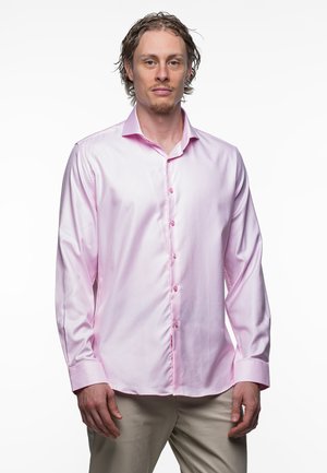 Man met licht krullend haar, gekleed in een lichtroze blouse met lange mouwen en beige broek, staand tegen een witte achtergrond.