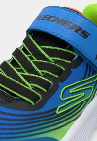 A Skechers sportcipő kék és zöld dizájnnal rendelkezik, texturált felsőrésszel, fekete fűzőkkel és egy kiemelkedő logóval egy pánton.