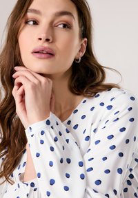 White blouse with blue polka dots, long sleeves with gathered cuffs, round neckline, featuring a button placket and soft fabric texture.