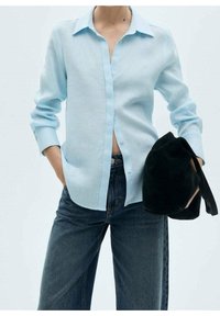 Chemise bleu clair à manches longues, à boutons avec une texture subtile, manches retroussées aux poignets, accompagnée d'un jean en denim foncé et d'un sac noir.