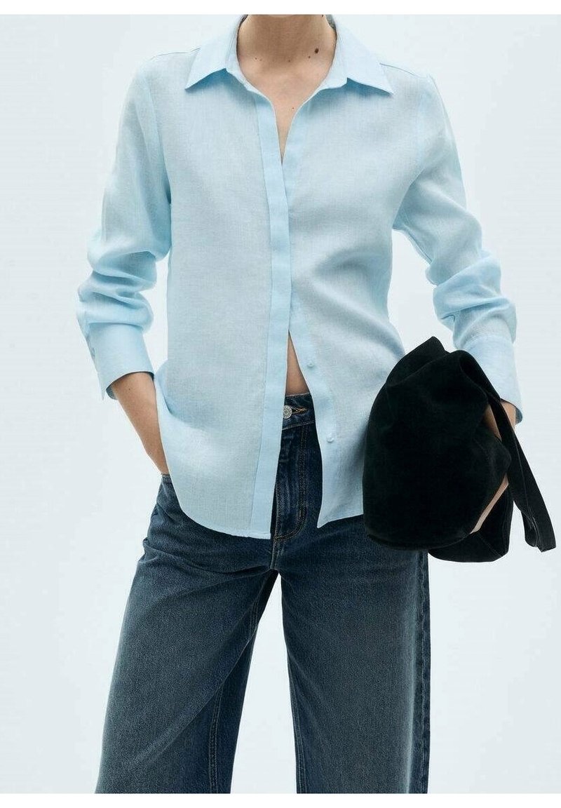 Chemise bleu clair à manches longues, à boutons avec une texture subtile, manches retroussées aux poignets, accompagnée d'un jean en denim foncé et d'un sac noir.