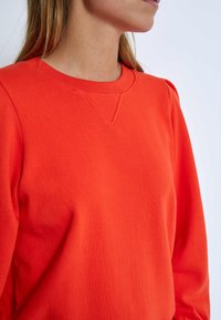 Red sweatshirt with a crew neckline and ribbed accents. Features soft texture and slightly puffed sleeves, emphasizing a modern design.