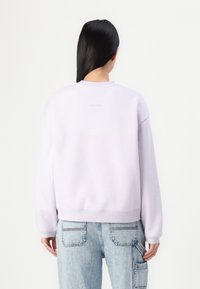 Sweatshirt violet deschis cu manșete și tiv țesute, având guler rotund și detalii subtile cu logo pe spate. Asortat cu blugi albaștri deschiși.