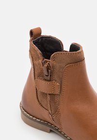 Brązowy skórzany botek sięgający do kostki z zamszowym panelem, bocznym zamkiem i pętelką do ściągania. Konstrukcja obejmuje detal w postaci przeszywanej podeszwy.