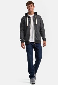 Haut de survêtement gris avec fermeture éclair et cordons de serrage, porté sur un t-shirt blanc, associé à un jean bleu foncé et des baskets grises. Look et style décontractés.