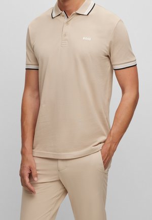 Polo beige à motif texturé, avec une patte de boutonnage à trois boutons, un col à rayures blanches et noires, et un petit logo sur la poitrine.