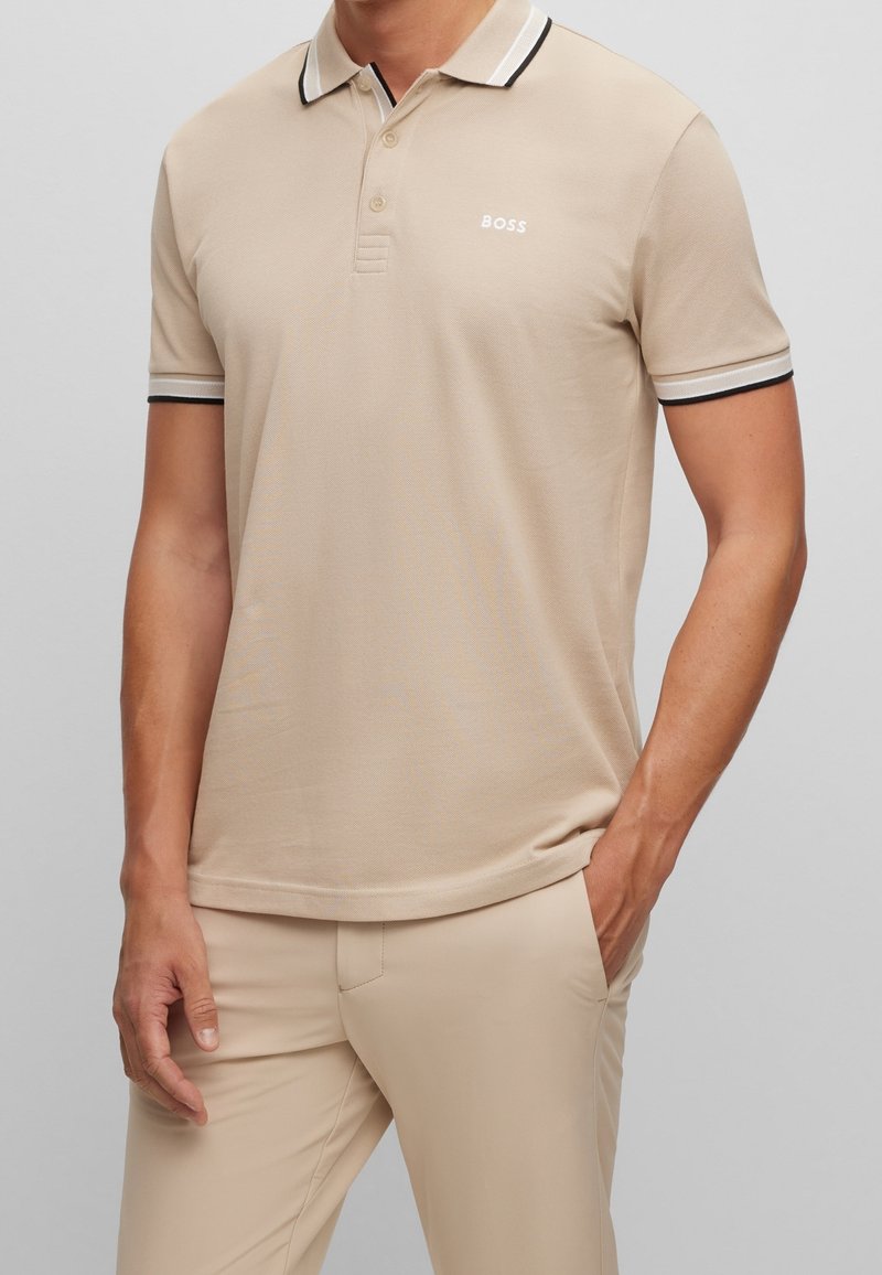 Polo beige à motif texturé, avec une patte de boutonnage à trois boutons, un col à rayures blanches et noires, et un petit logo sur la poitrine.