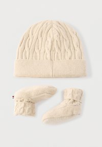 Flødefarvet strikket babyhue med kabelmønstre og matchende bløde futter på en lys baggrund.