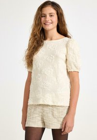 Kreeminen, tekstuurilla varustettu pusero, jossa on puffihihat ja kukkakuosi. Yhdistetty vaaleisiin kudottuihin shortseihin. Taustana on neutraali valkoinen.