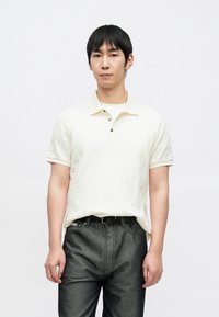 Hellbeiges Poloshirt mit strukturiertem Rautenmuster, ausgestattet mit einem klassischen Kragen und zwei Knöpfen. Kombiniert mit dunkelgrauen Jeans.