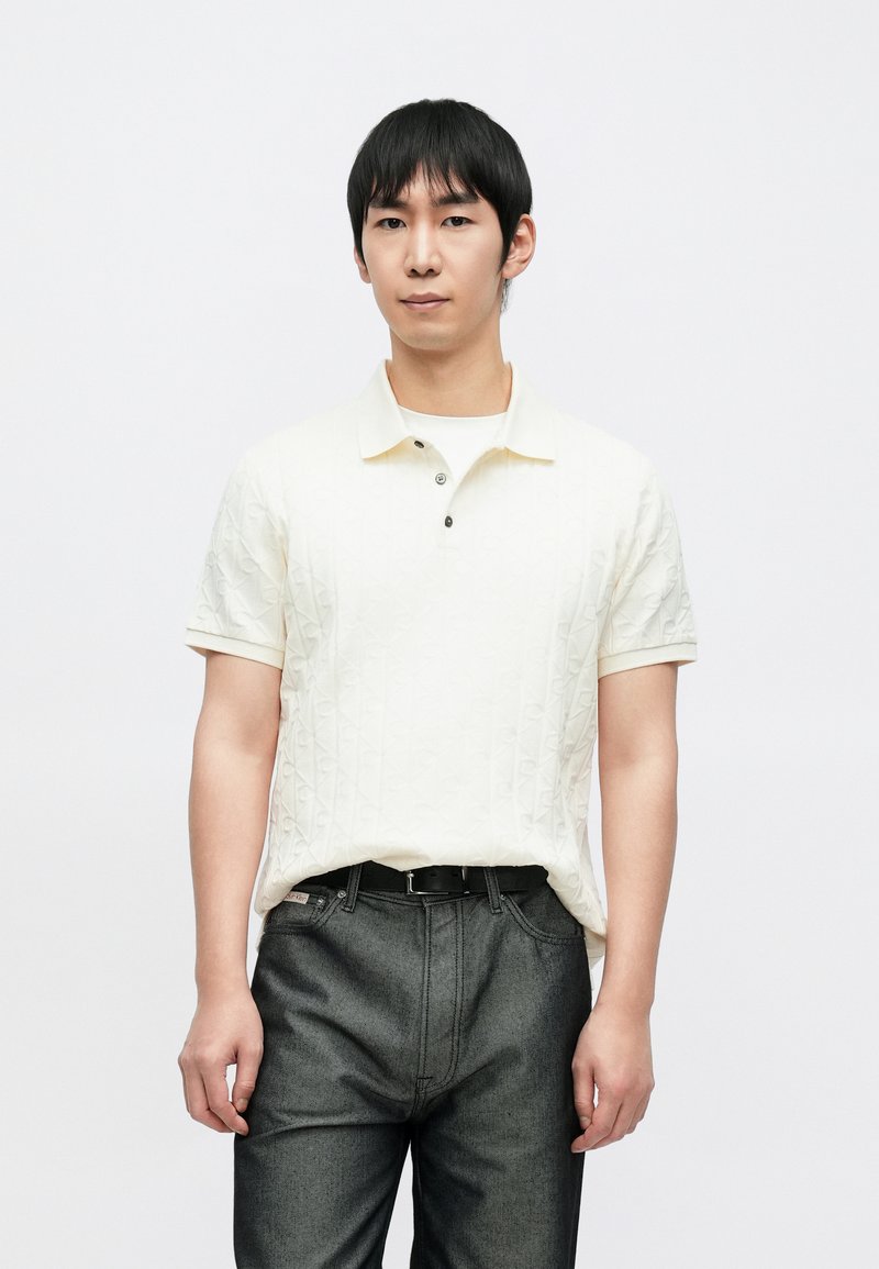 Hellbeiges Poloshirt mit strukturiertem Rautenmuster, ausgestattet mit einem klassischen Kragen und zwei Knöpfen. Kombiniert mit dunkelgrauen Jeans.