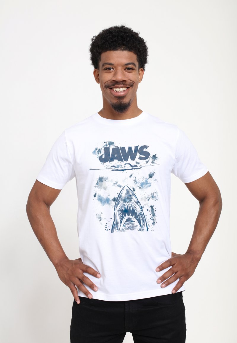 Bílé bavlněné tričko s modrým grafickým potiskem žraloka a nápisem "JAWS" nahoře, s akcenty ve formě skvrn v různých odstínech modré.