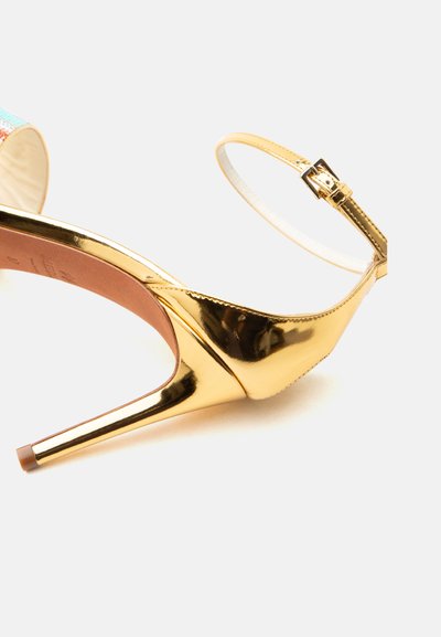 Złota metaliczna sandałka na szpilce z cienkim paskiem wokół kostki i klamrą, widok z boku i z tyłu na białym tle.