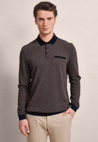 Langärmliges Poloshirt aus dunkelgrauem Stoff mit strukturiertem Muster, mit einem marinefarbenen Kragen und einer Tasche mit Knopfdetail, kombiniert mit beigen Hosen.