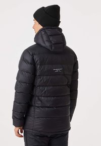 Svart puffjacka med huva, med horisontell quiltning och en logotyp på ryggen. Slät textur och figursydd design.