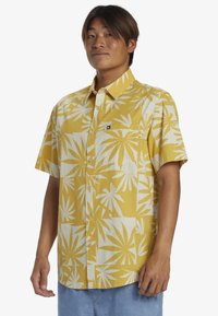 Camicia a maniche corte in tessuto giallo con motivi a foglia bianchi, con bottoni sul davanti e una tasca sul petto con un piccolo etichetta logo.