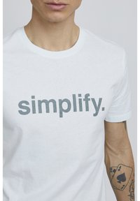 Hvid bomulds t-shirt med ordet "simplify." i store grå bogstaver. Pasformen er standard med rund hals og korte ærmer.