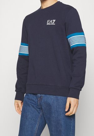 Mężczyzna ubrany w granatową bluzę z niebiesko-białymi paskami na rękawach i białym logo EA7 Emporio Armani na piersi.