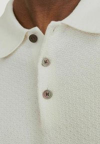 Polo bianco con texture e finitura morbida a maglia, dotato di colletto a punta e tre bottoni rotondi marroni sulla patta.
