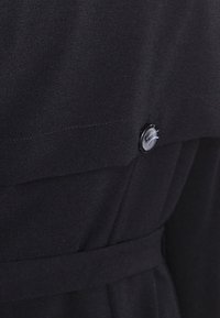 Schwarzer Stoff mit einem einzelnen schwarzen Knopf, der in einer Naht in der Nähe eines Gürtels oder eines Taillenbunds an einem Kleidungsstück angenäht ist.